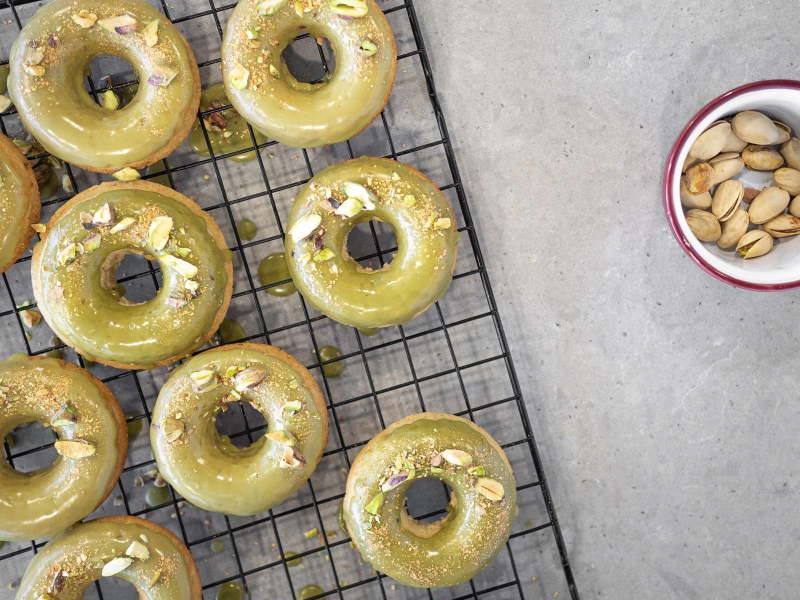 Udekorowane zielonym lukrem z matchą, pistacjami i złotymi drobinkami pieczone donuty bez mleka i jajek na kratce do studzenia.