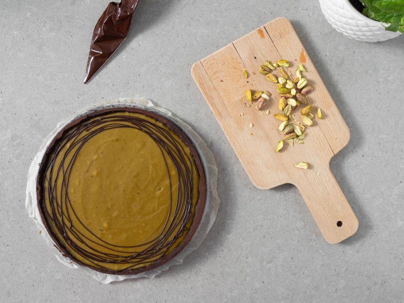 Tarta pistacjowa udekorowana rozpuszczoną czekoladą. Obok na deseczce posiekane pistacje.