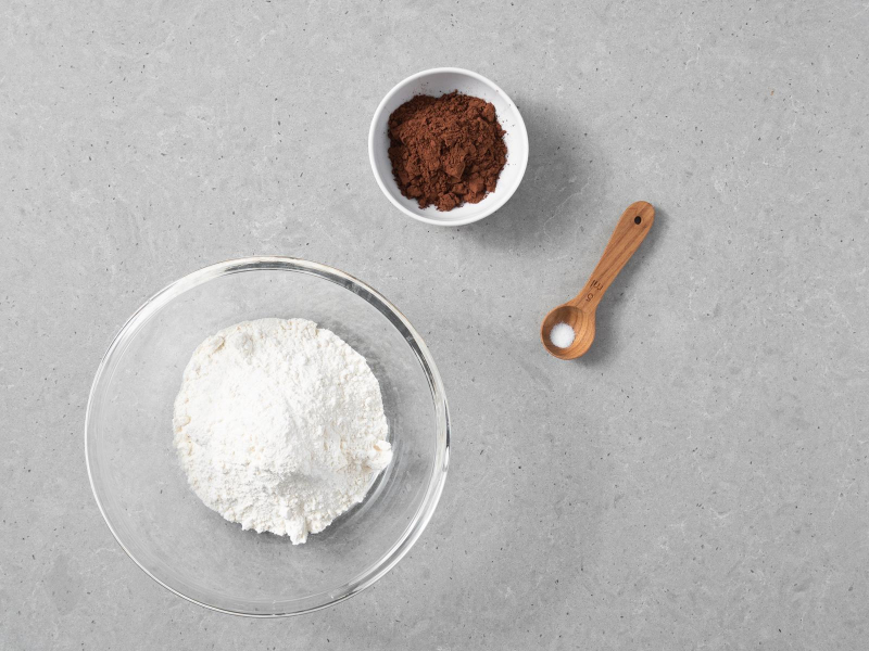 Mąka pszenna w szklanej misce. Obok ciemne kakao w miseczce i sól na drewnianej łyżeczce.