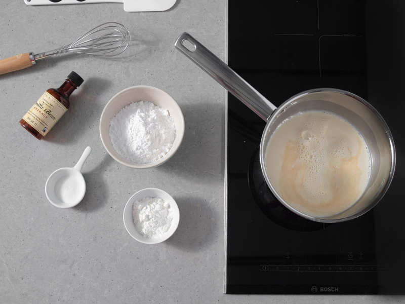 W garnku na kuchence napój sojowy z dolanym ekstraktem waniliowym. Obok miseczki cukrem pudrem,  skrobią kukurydzianą i agarem oraz opakowanie ekstraktu waniliowego taylor and colledge