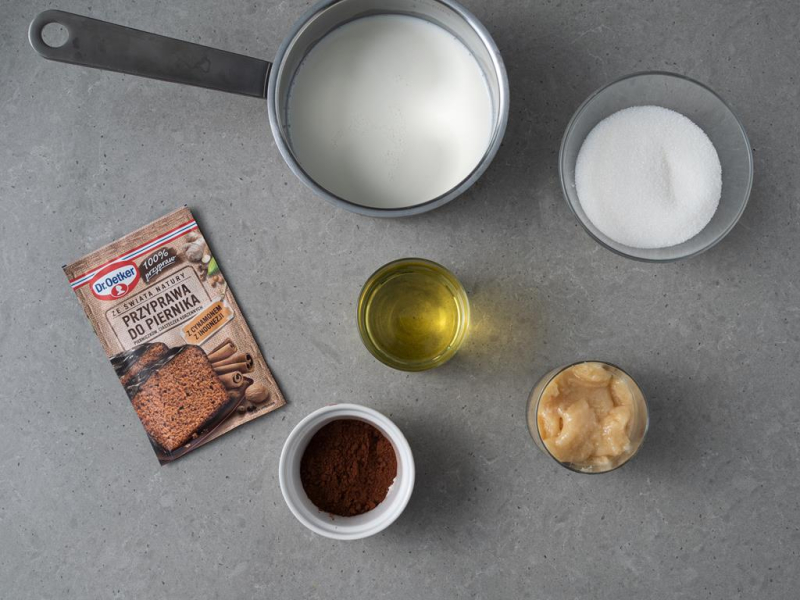 W garnku mleko. Obok cukier w miseczce, olej, miód i kakao. Na blacie opakowanie z przyprawą korzenną Dr. Oetkera.