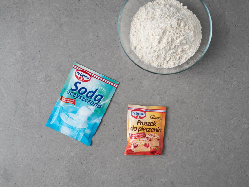W dużej misce mąka pszenna. Obok opakowanie sody oczyszczonej oraz proszku do pieczenia Dr. Oetkera.