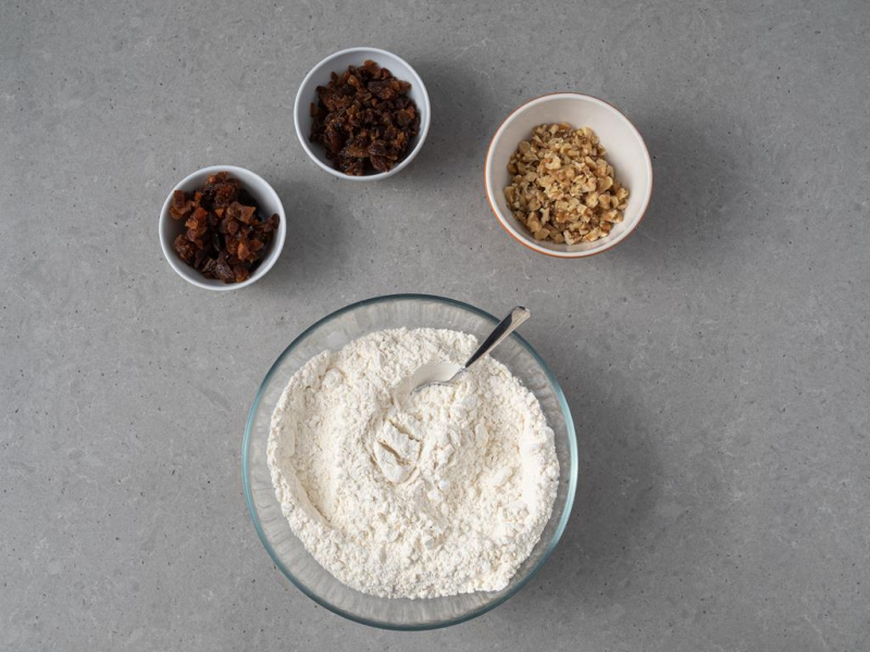 W misce mąka pszenna wymieszana z proszkiem do pieczenia i sodą. Obok w miseczkach pokrojone bakalie.