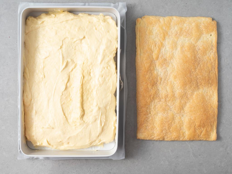 Krem budyniowo-śmietankowy przełożony na blat ciasta francuskiego. Obok sam blat ciasta.