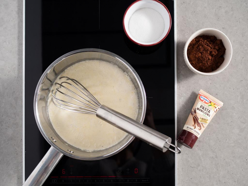 śmietanka w rondelku na palniku kuchenki, obok cukier i kakao w osobnych miseczkach oraz opakowanie pasty waniliowej intense od dr. oetkera