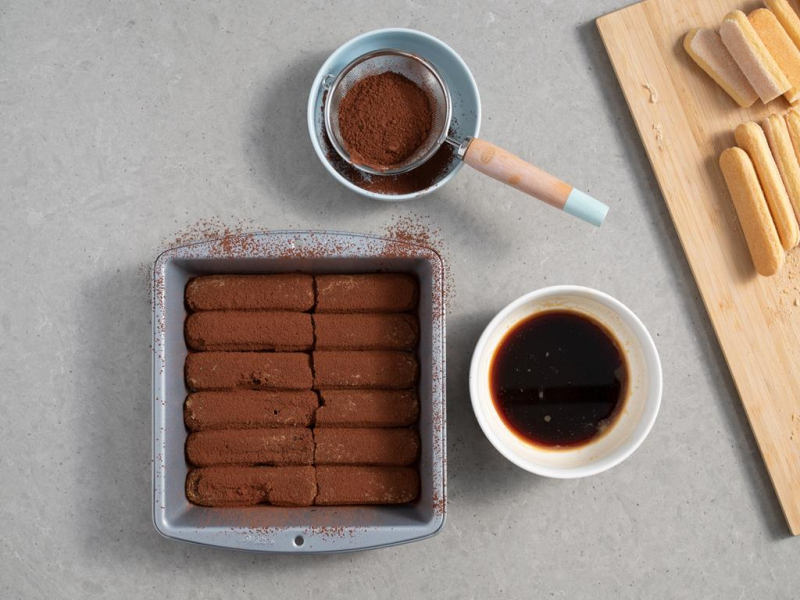 biszkopty nasączone kawą i oprószone kakao w kwadratowej formie do pieczenia marki Wilton, obok miseczka z kawą i sitko z ciemnym kakao od dr. oetkera