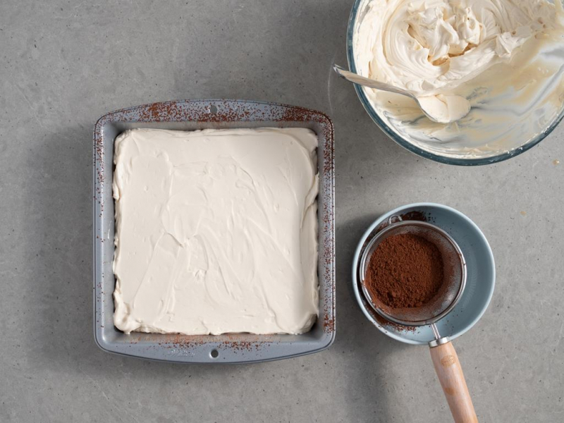 pierwsza warstwa kremu baileys wyłożona na biszkoptach, obok krem w misce i sitko z kakao