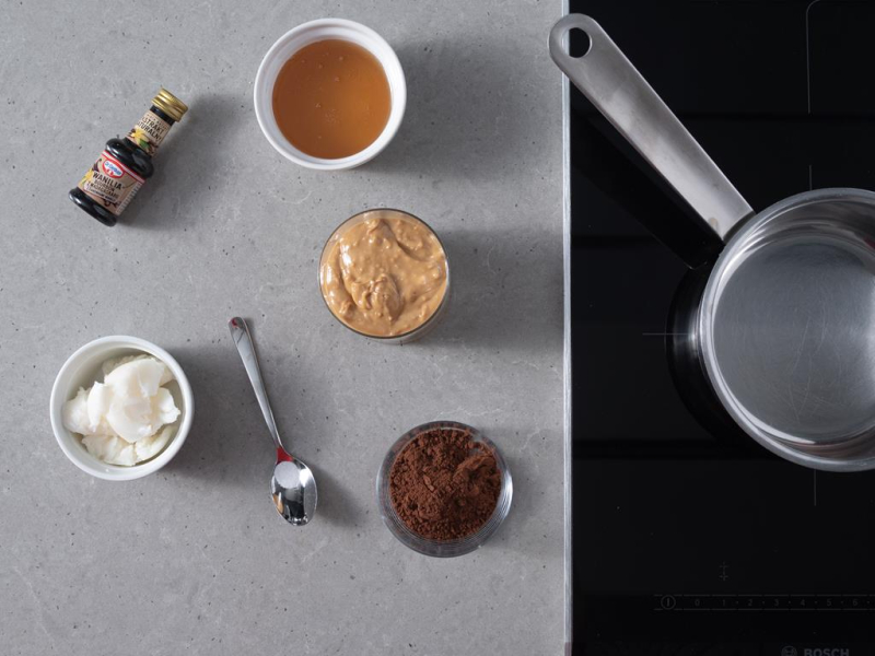 Garnek na płycie indukcyjnej firmy Bosch. Obok w miseczce kakao ciemne, masło orzechowe, miód, szczypta soli na łyżeczce, ekstrakt z wanilii Dr. Oetkera i olej kokosowy.