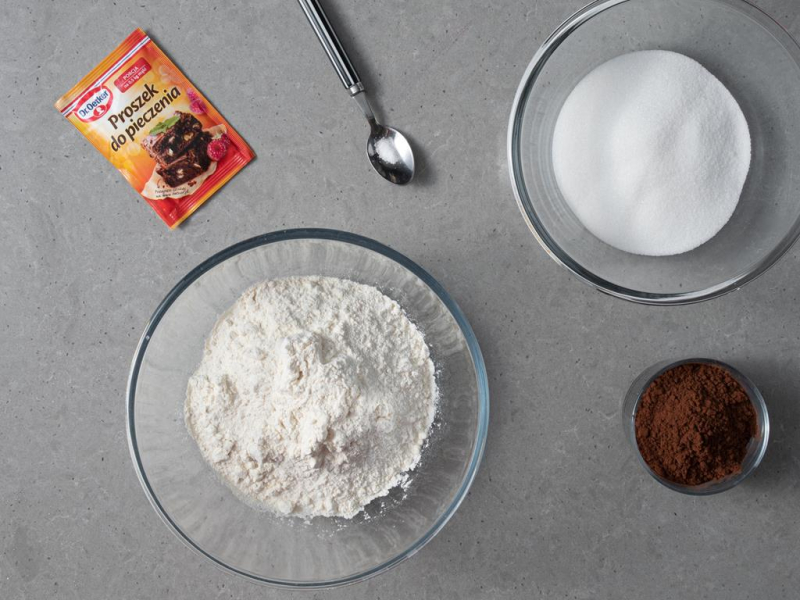 Mąka w dużej, szklanej misie. Obok cukier oraz ciemne kakao. Na łyżeczce odrobina soli. A obok opakowanie proszku do pieczenia Dr. Oetkera.