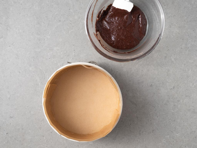 W szklanej misie wymieszane składniki na ciasto czekoladowe. Obok tortownica wyłożona papierem do pieczenia.