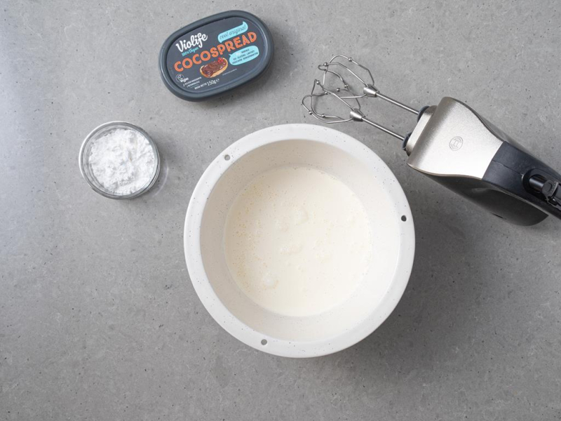 W misie schłodzona śmietanka roślinna, obok cukier puder oraz czekoladowy serek roślinny.