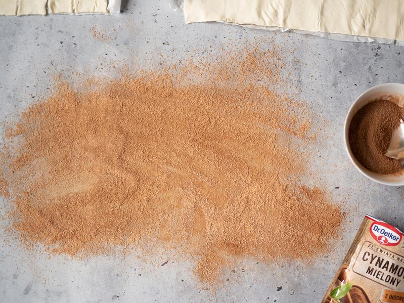 na blacie kuchennym rozsypany cukier z cynamonem, obok dwa arkusze ciasta francuskiego na palmiery, opakowanie cynamonu mielonego dr. oetkera oraz miseczka z cukrem wymieszanym z cynamonem
