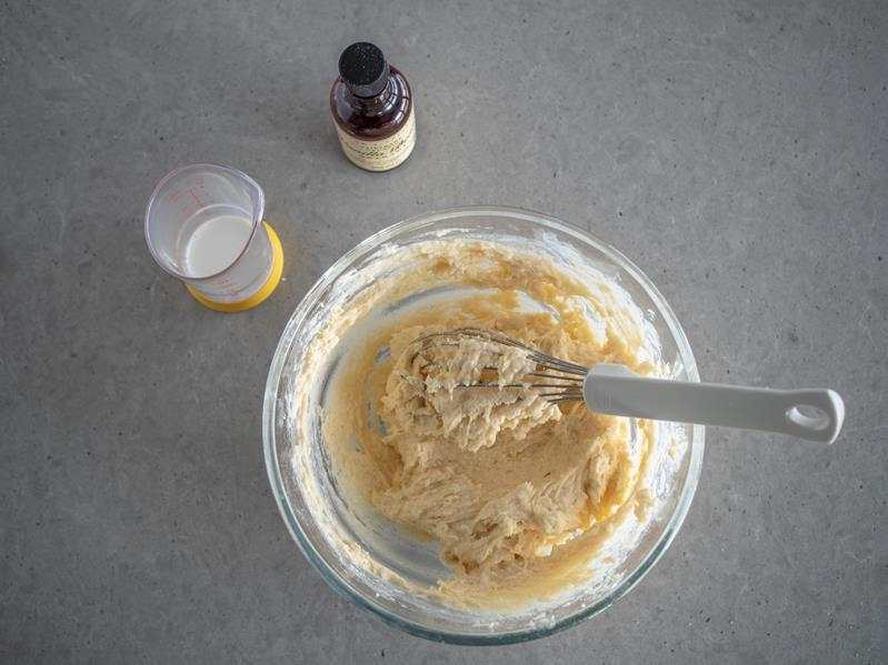 szklana miska z ciastem na pączki z dodatkiem sera typu włoskiego, rózga kuchenna, opakowanie ekstraktu waniliowego taylor & colledge, szklana miarka kuchenna z mlekiem