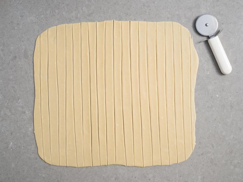 rozwałkowane ciasto na rurki z kremem pocięte na paski, obok radełko