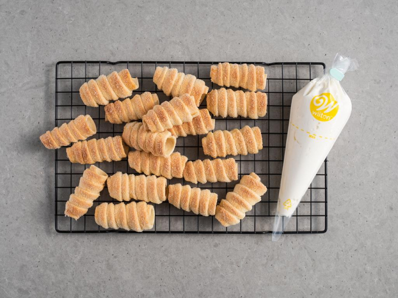 upieczone rurki na kratce do studzenia, obok krem waniliowy w rękawie cukierniczym Wilton
