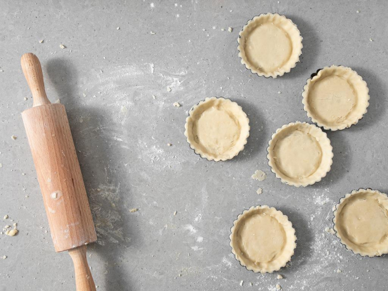 Wyłożone ciastem kruchym foremki do tartaletek. Obok wałek do ciasta.