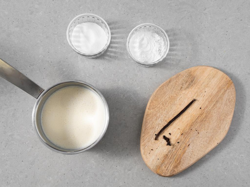 Laska wanilii na drewnianej deseczce. Obok napój sojowy w rondelku oraz cukier i skrobia kukurydziana w osobnych miseczkach