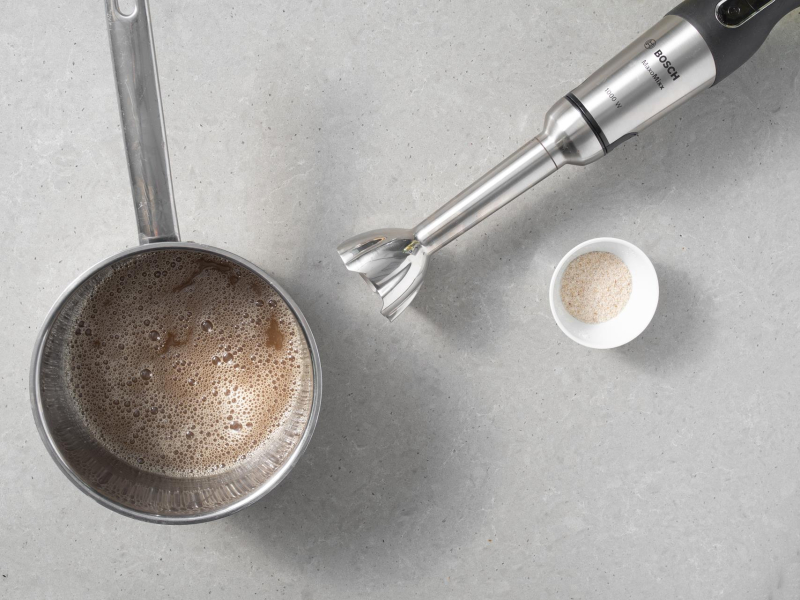 W garnku rozpuszczony cukier z pastą waniliową i napojem migdałowym. Obok w miseczce babka płesznik i blender ręczny marki Bosch.