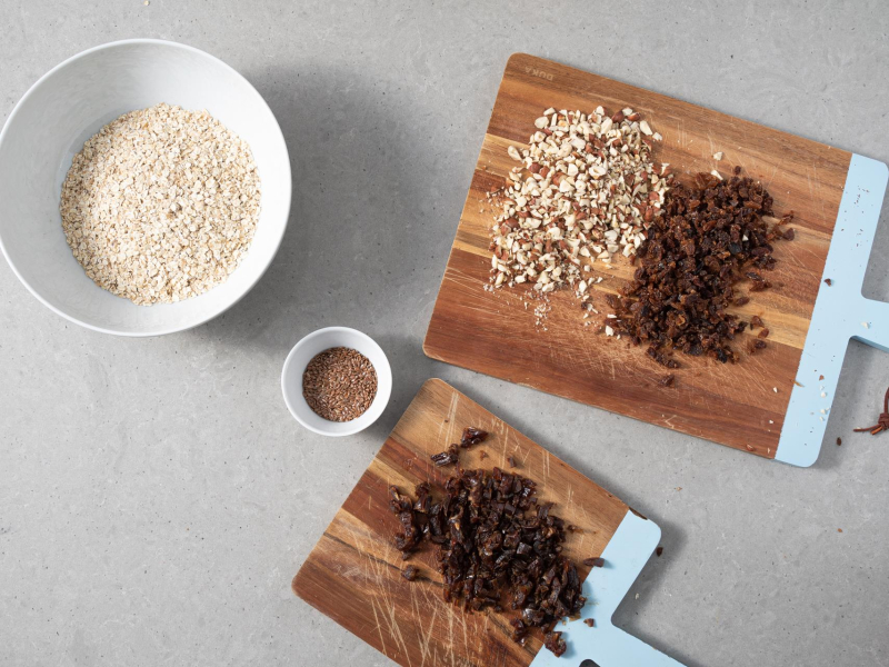 W misie płatki owsiane błyskawiczne. Obok w miseczce siemię lniane. Na deskach pokrojone bakalie.