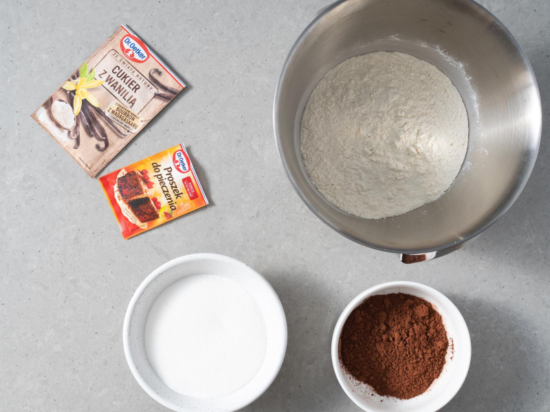 W misie robota kuchennego m膮ka pszenna. Obok w miseczce kakao i cukier. A na blacie opakowanie cukru z wanili膮 i proszek do pieczenia.