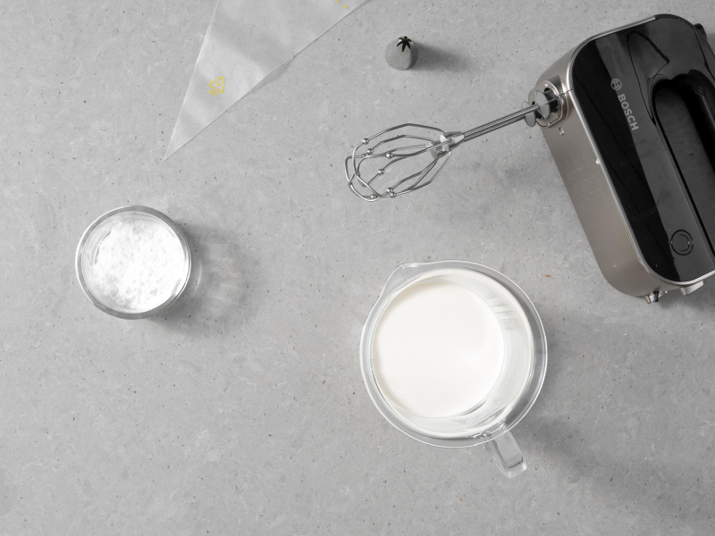 Na blacie mikser r臋czny, obok cukier puder w miseczce, r臋kaw cukierniczy, tylka i 艣mietanka ro艣linna.