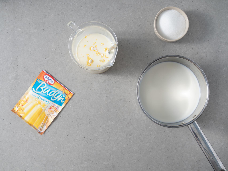 Na blacie opakowanie budyniu o smaku waniliowym Dr. Oetkera, obok cukier z miseczce. W garnku mleko i obok w wysokim naczyniu mleko z proszkiem budyniowym.