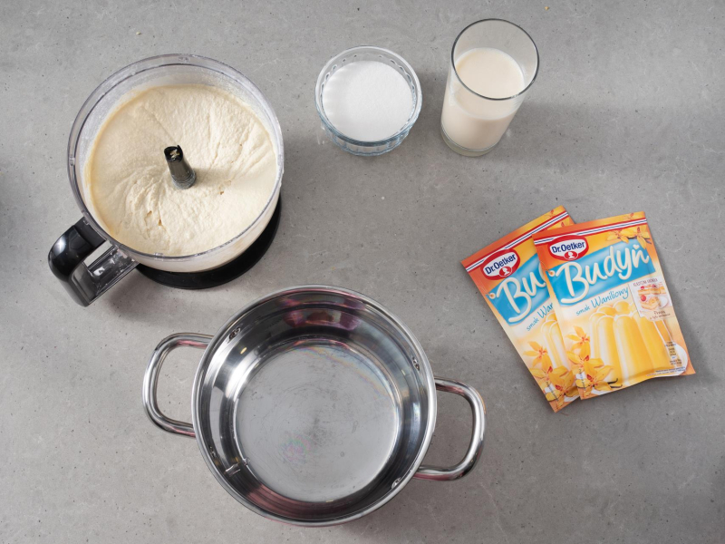 W misie malaksera masa z tofu i soku z cytryny. Obok garnek, mleczko kokosowe, napój sojowy w szklance. Cukier w miseczce i dwa opakowania budyniu o smaku waniliowym Dr. Oetkera.