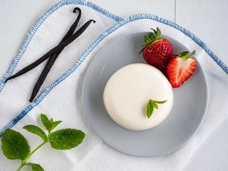 panna cotta na talerzyku, obok świeże truskawki, gałązka mięty i laski wanilii