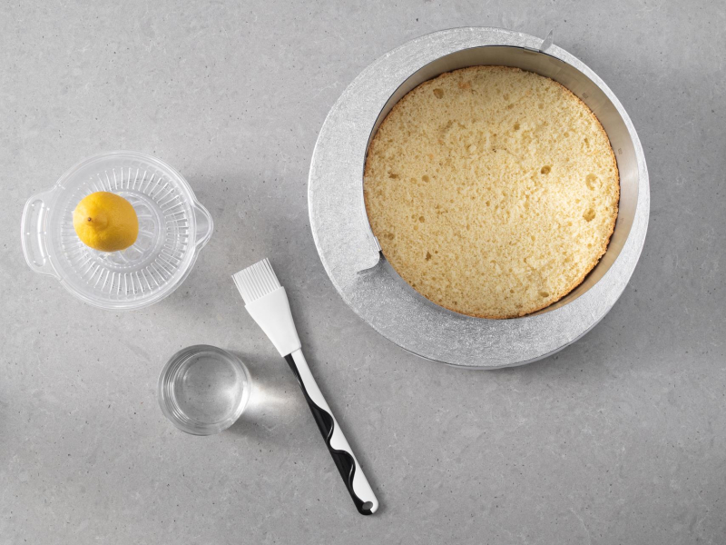 Blat biszkoptu w rancie cukiernicznym. Obok pędzelek silikonowy i składniki na poncz.