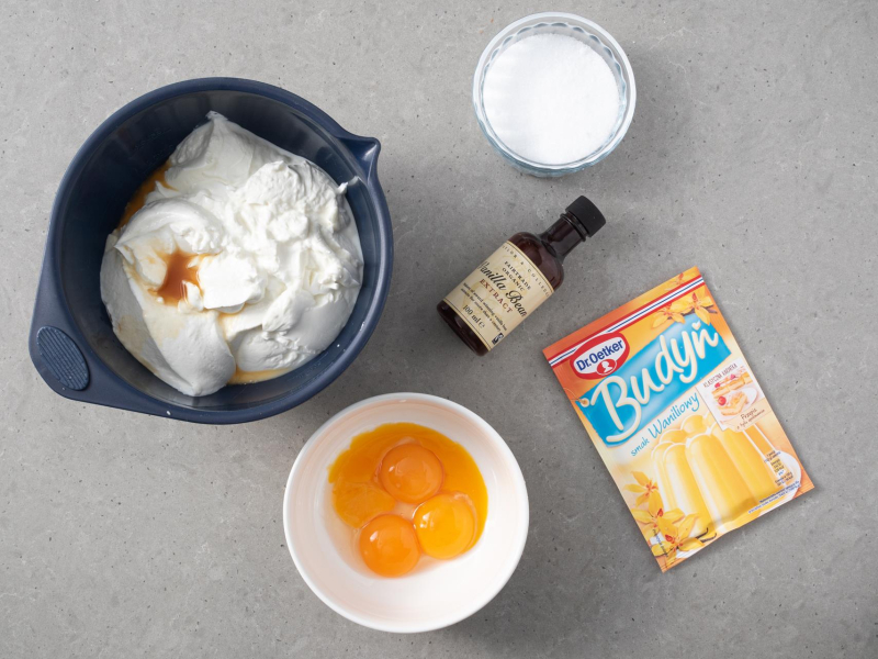 W granatowej misce skyr, proszek do pieczenia i ekstrakt waniliowy. Obok budyń o smaku waniliowym Dr. Oetkera, ksylitol i ekstrakt waniliowy.