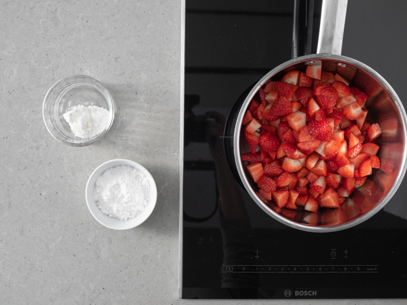Pokrojone truskawki w garnku. Obok w miseczce cukier i skrobia kukurydziana.