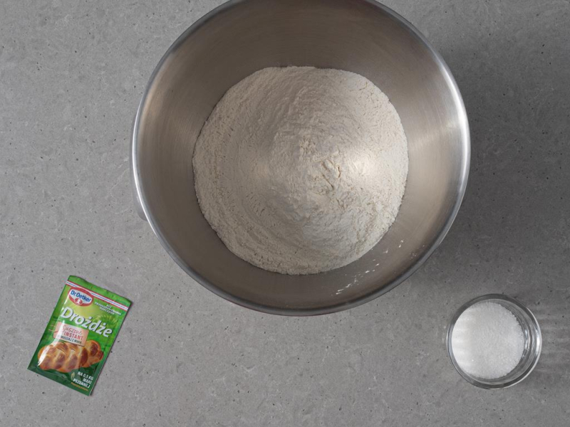 Mąka pszenna w misie miksera firmy Bosch. Obok drożdże suszone Dr. Oetker oraz cukier w miseczce.