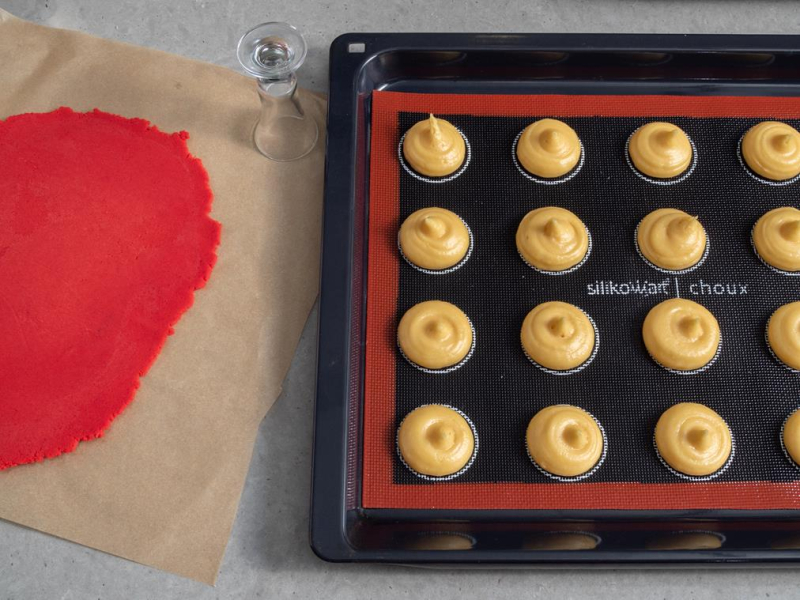 w blaszce na macie silikonowej wyszprycowane nieupieczone ptysie obok czerwone rozwałkowane ciasto kruche