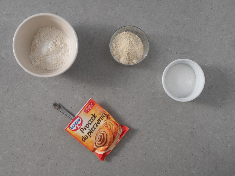 W miskach: mąka pszenna, migdałowa oraz cukier. Na blacie opakowanie proszku do pieczenia Dr. Oetkera.