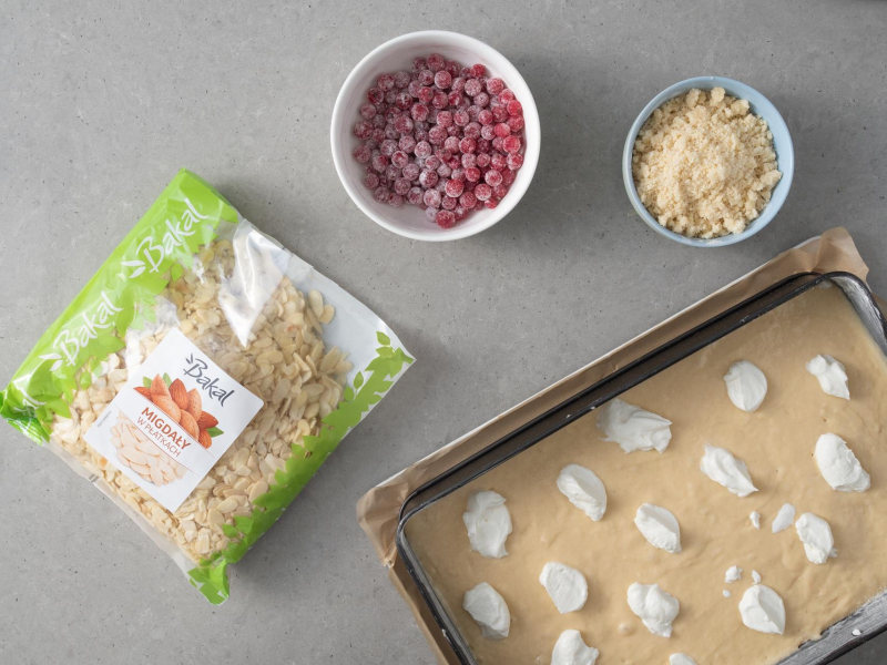 W blaszce wyłożone surowe ciasto z kleksami twarogu. Obok w miskach: kruszonka, czerwone porzeczki i płatki migdałów.