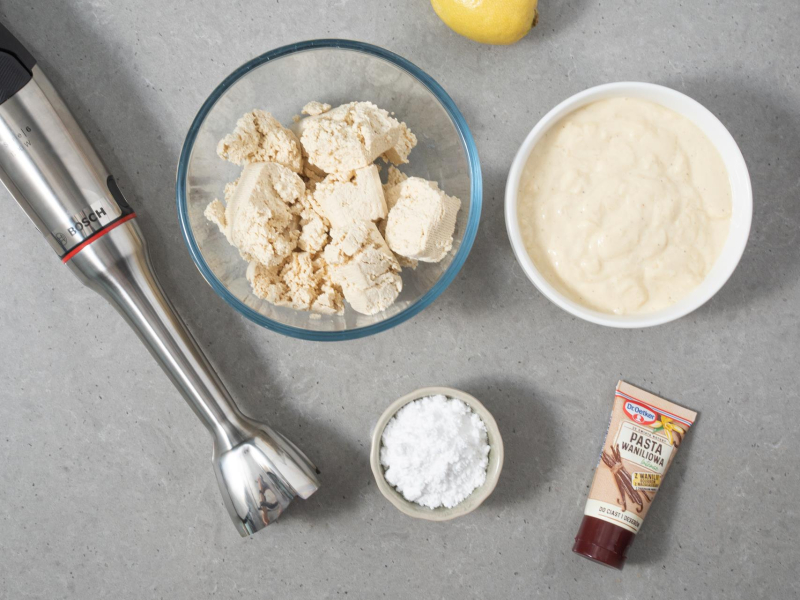 Na blacie w dużej misce pokruszone tofu. Obok jogurt sojowy, cukier puder, pasta waniliowa, cytryna i blender ręczny firmy Bosch.