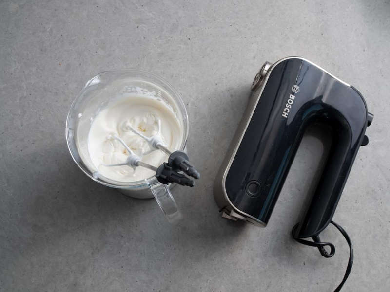 w szklanym naczyniu ubita śmietanka obok ręczny mikser bosch