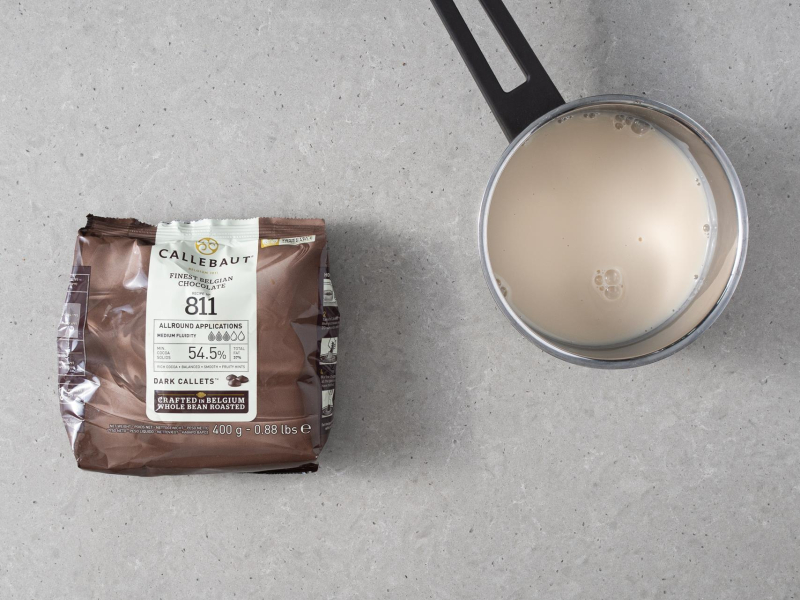 Na blacie kuchennym opakowanie czekolady ciemnej Callebaut 811 obok napój migdałowy w garnuszku.