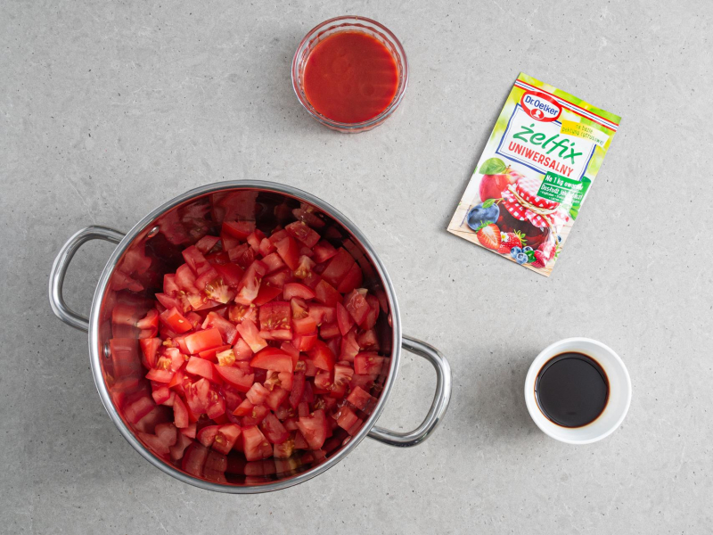 W garnku pokrojone pomidory. Obok na blacie opakowanie żelfixu uniwersalnego, ocet balsamiczny i koncentrat pomidorowy.