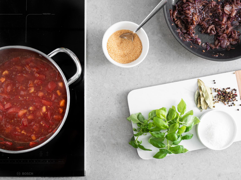 W garnku pomidory z żelfixem, cukrem oraz octem balsamicznym. Na desce świeże zioła. Na patelni skarmelizowana cebula i w małej miseczce cukier trzcinowy.