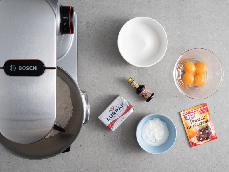 Na blacie kuchennym robot kuchenny firmy Bosch. Obok kostka masła lurpak, opakowanie proszku do pieczenia, ekstraktu waniliowego, żółtka w miseczce, cukier i kwaśna śmietana.