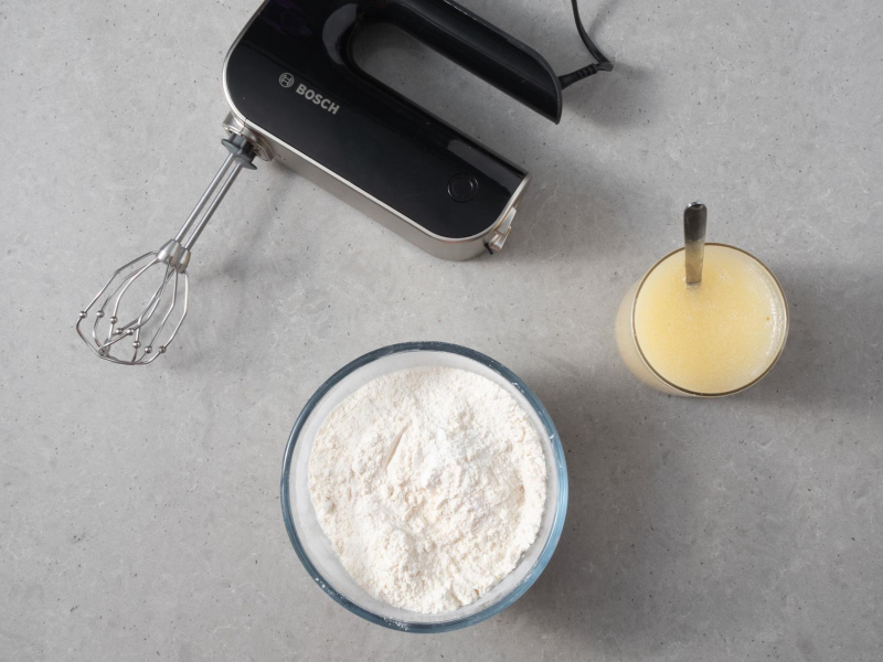 Na blacie kuchennym miska z suchymi składnikami do ciasta i dzbanek z mokrymi. Obok mikser ręczny firmy Bosch.