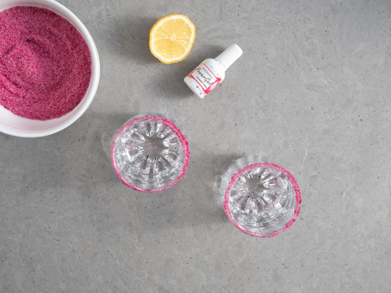 W miseczce zabarwiony cukier. Obok szklanki z kolorowym cukrem.