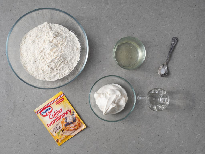 W dużej misie mąka. Obok w miseczce wegańska śmietana i olej. Na łyżeczce sól i w kieliszku spirytus. Na blacie opakowanie cukru wanilinowego Dr. Oetkera.