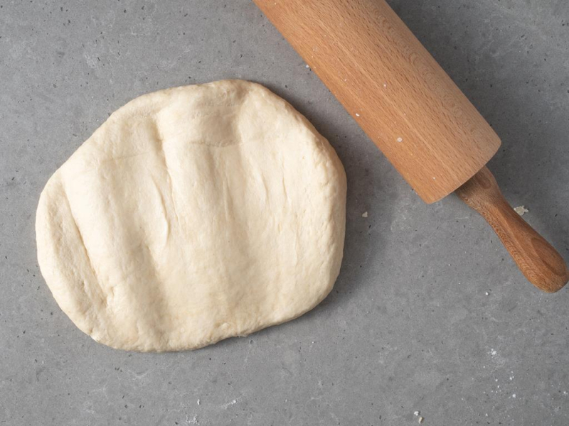 Przygotowane ciasto na faworki wegańskie. Obok drewniany wałek do pieczenia.