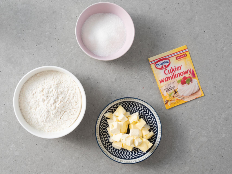 Na blacie kuchennym w miseczkach pokrojone zimne masło, mąka oraz cukier. Obok opakowanie cukru waniliowego Dr. Oetkera.