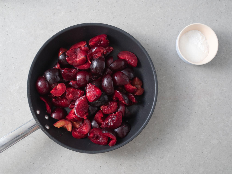Na patelni pokrojone śliwki. Obok miseczka z cukrem i mąką ziemniaczaną.