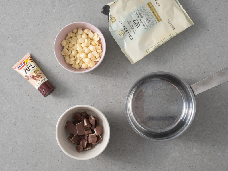 Na blacie kuchennym biała czekolada, czekoladki kinder oraz pasta z wanilii.