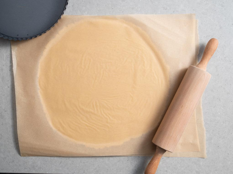 rozwałkowane ciasto pomiędzy arkuszami papieru do pieczenia obok wałek drewniany