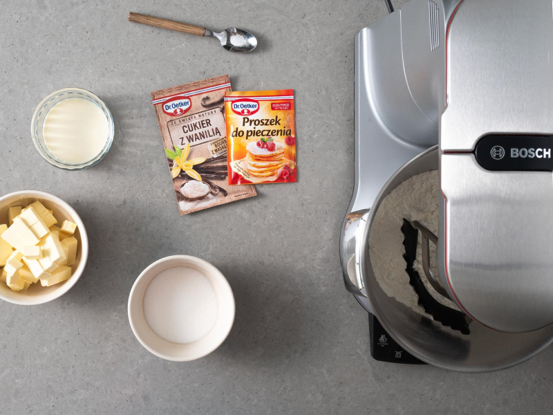 Na blacie robot kuchenny firmy Bosch. Obok cukier z wanilią Dr Oetker, proszek do pieczenia, łyżeczka z solą, schłodzona margaryna, napój sojowy i cukier w miseczce.